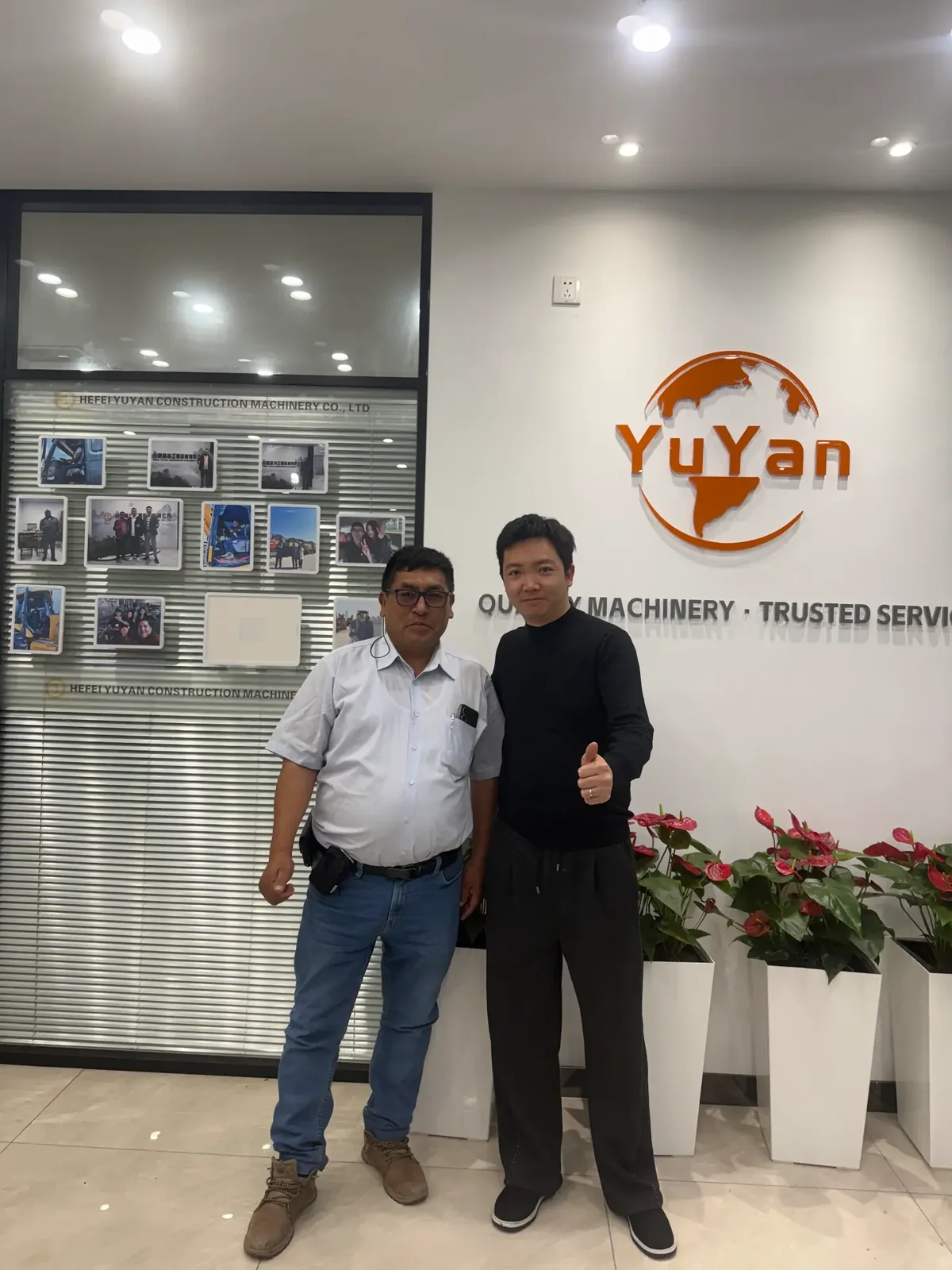 Two people giving thumbs-up while posing in front of the YUYAN-brand wall in the office